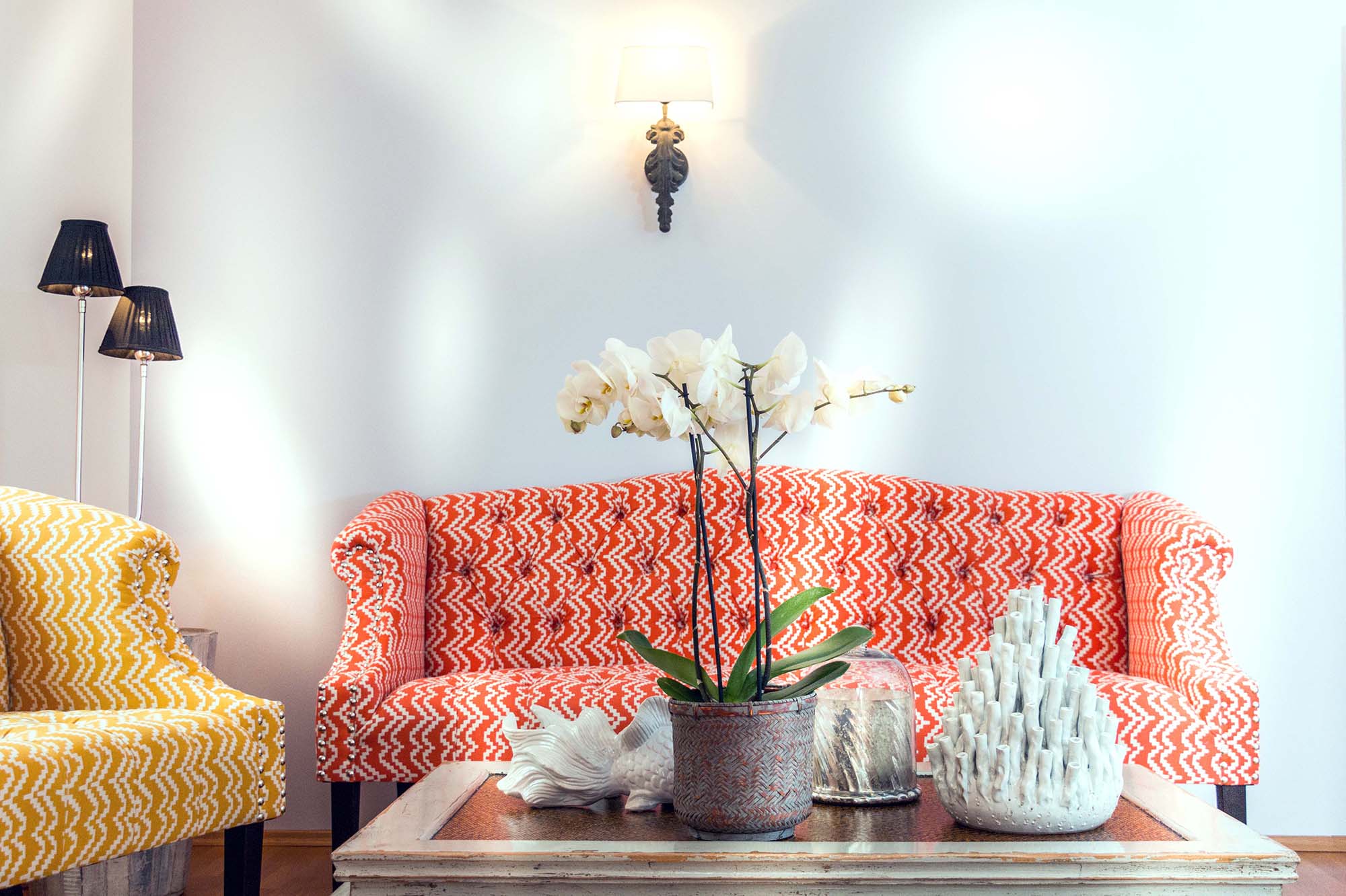 Orange-weiß gemustertes Sofa und gelb-weiß gemusterter Sessel mit einem Sofatisch aus Holz und Glas, darauf maritime Dekoration in Weiß.