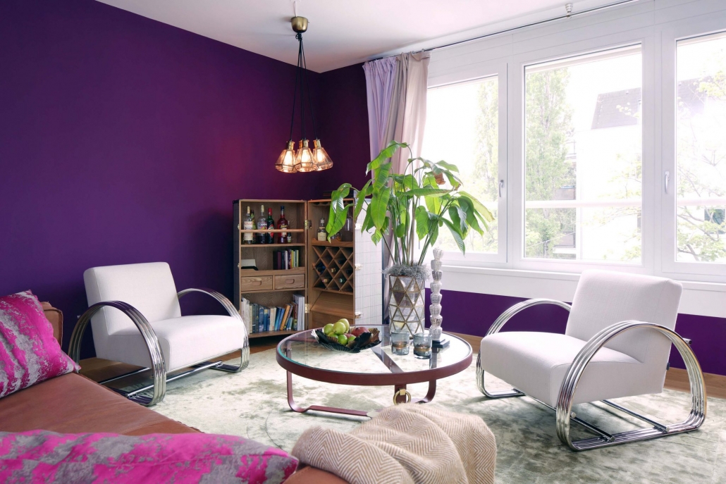 Wohnraum eingerichtet mit violetten Wänden, weißen Lounge-Sesseln, rundem Sofatisch aus Glas, Kofferbar und Ledersofa.