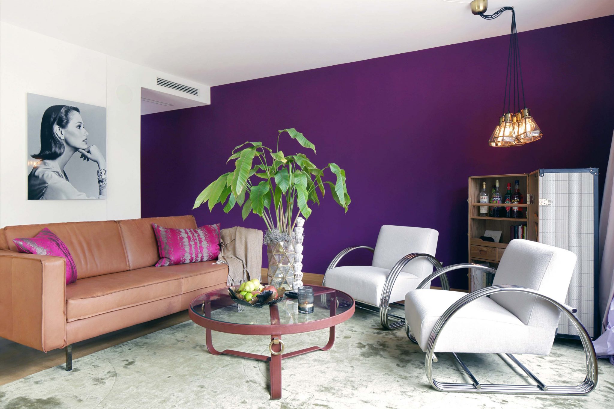 Wohnraum eingerichtet mit violetten Wänden, weißen Lounge-Sesseln, rundem Sofatisch aus Glas, Kofferbar, Ledersofa und Portrait an der Wand.