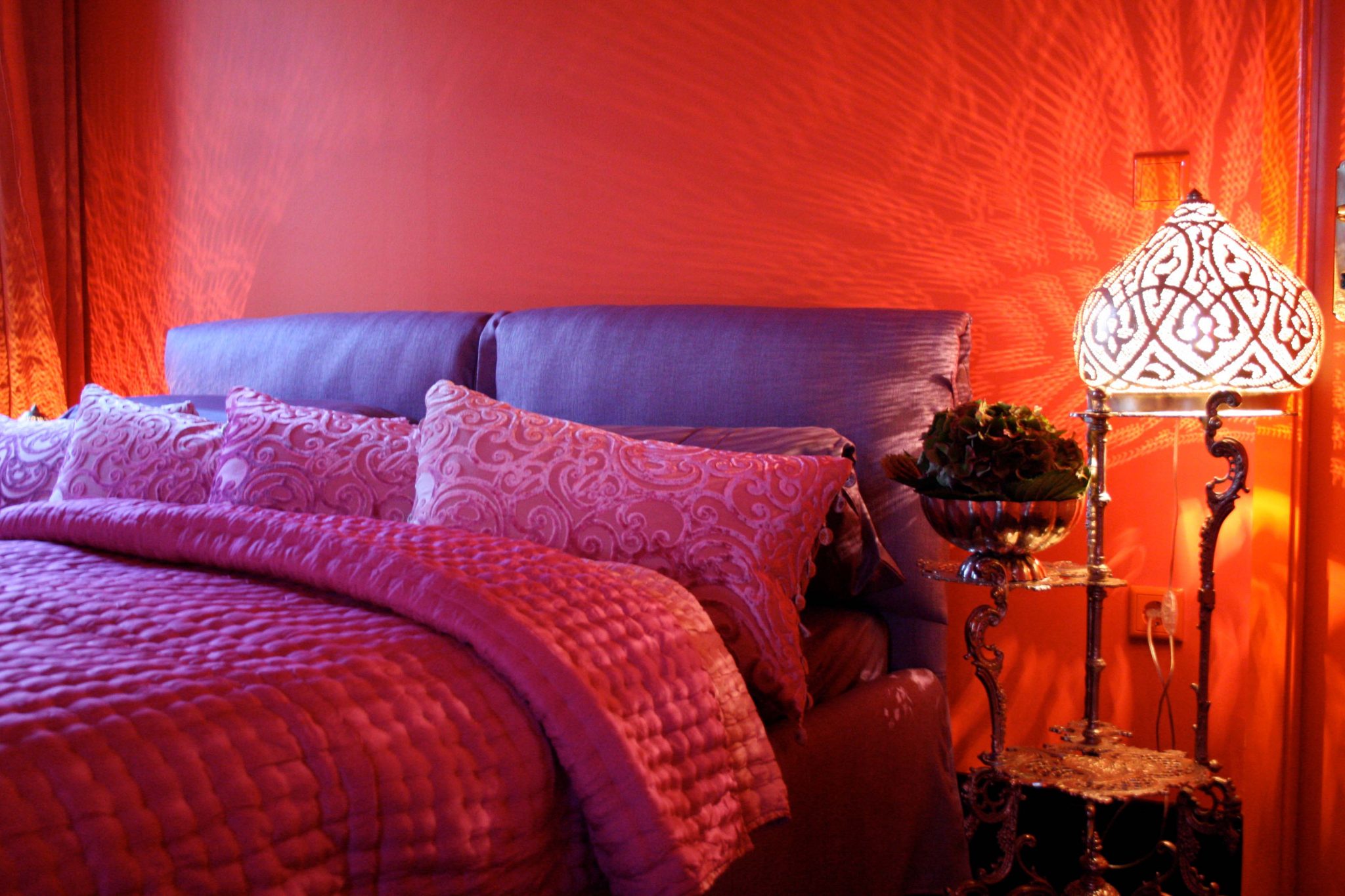 Orange gestrichenes Zimmer, violett gepolstertes Doppelbett mit magentafarbener Decke und Kissen, daneben orientalischer Nachttisch mit Lampe in Zwiebelform.