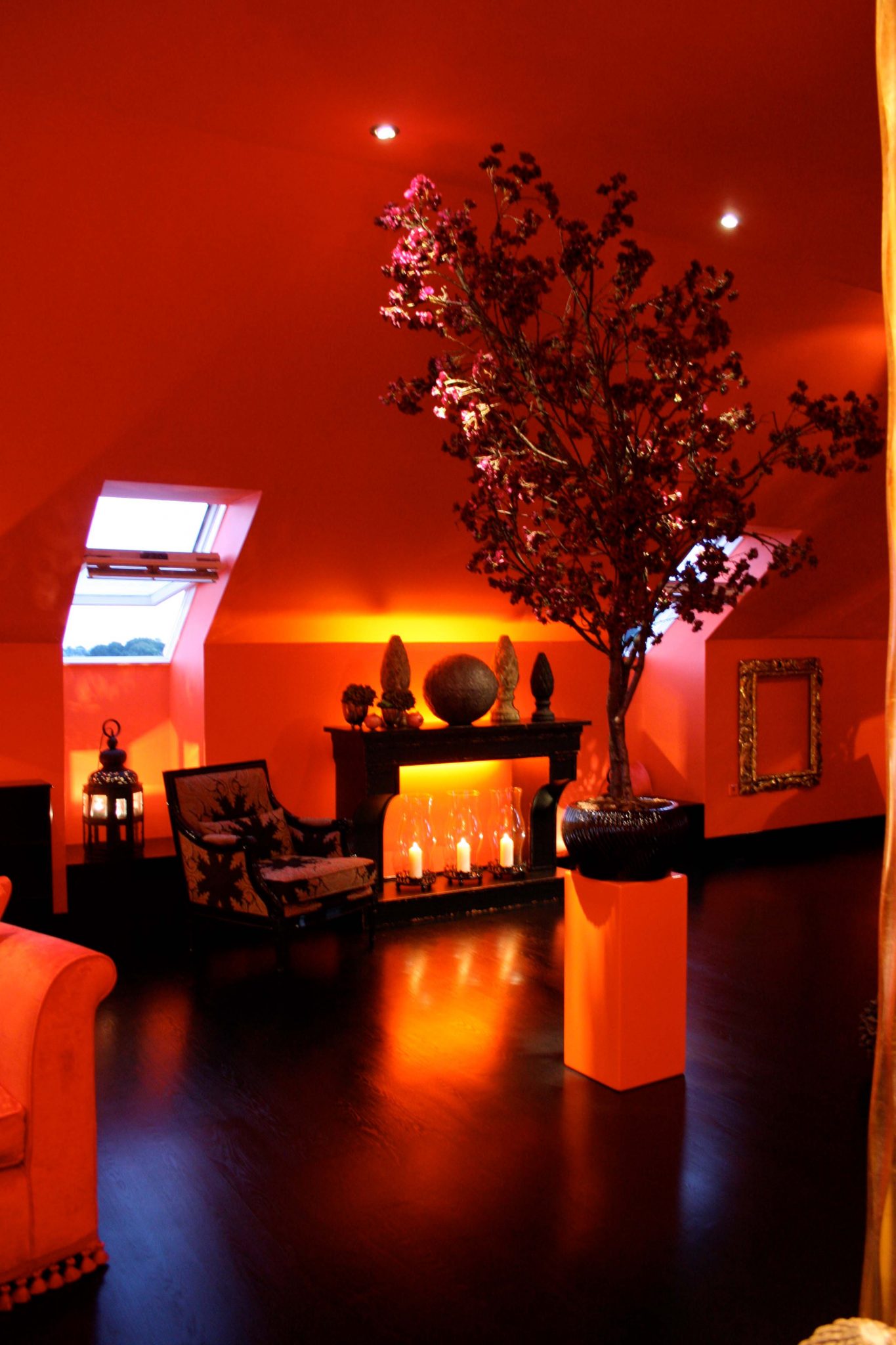 Lounge-Sessel mit gemustertem Stoffbezug, beleuchtetem Sideboard mit Deko und kleinem Baum in der Raummitte, Zimmer orange gestrichen.