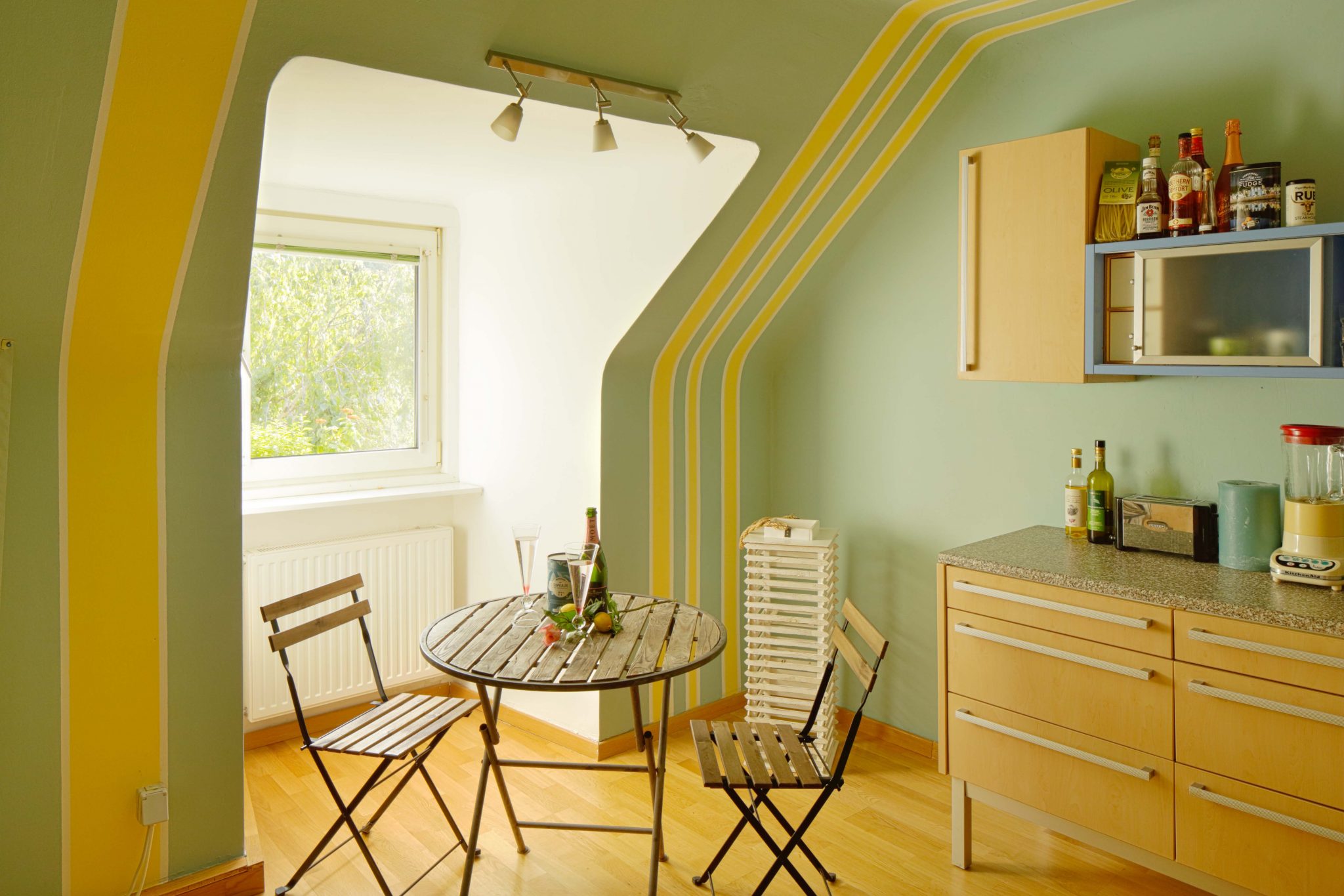 Mittels mit Streifen bestrichenen Wänden inszenierte Dachschräge in Gelb und Hellgrün, kleiner runder Tisch mit zwei kleinen Stühlen als Interior.