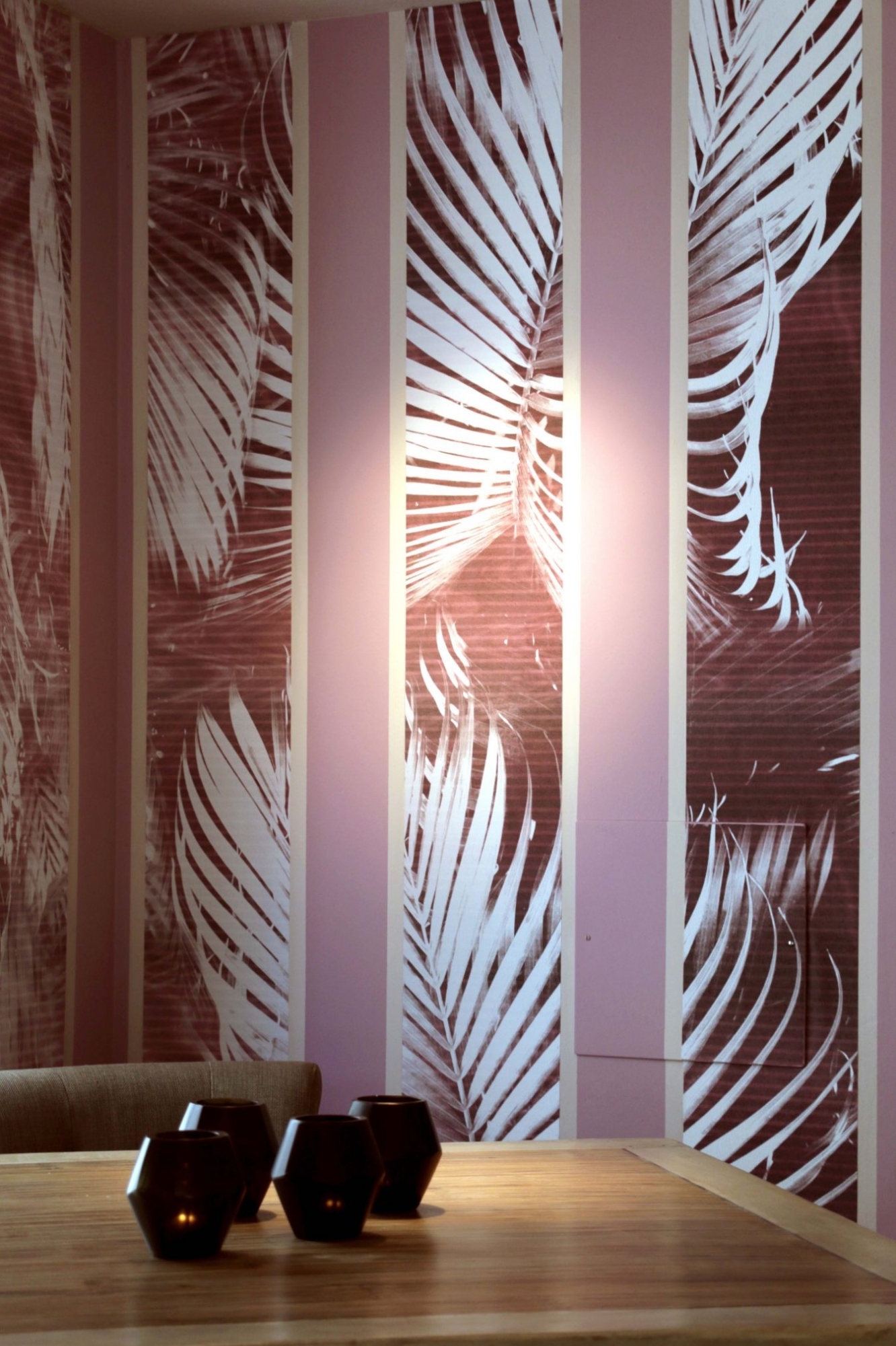 Gestreift gestaltete Wand mit Tapete mit Palmenblättern-Motiv und Weiß-Altrosa-Weiß gemalten Farbstreifen, davor ein Holztisch mit Teelichtern.