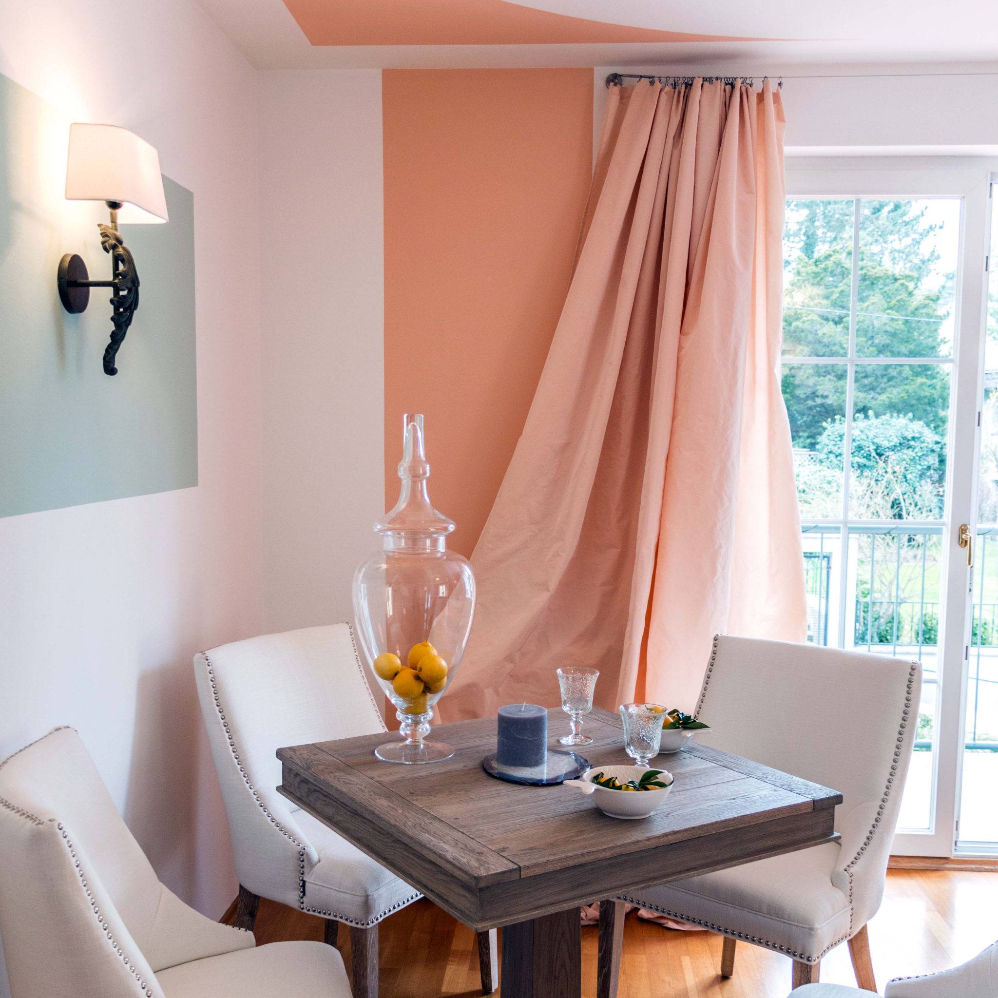 Quadratischer Holztisch vor aprikot-farbenem Vorhang mit Blick auf den Balkon.