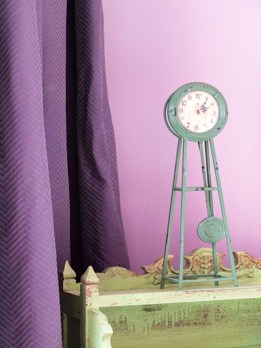 Violettes Zimmer mit passendem Vorhang und grüner, kleiner Standuhr auf grünem Sideboard.