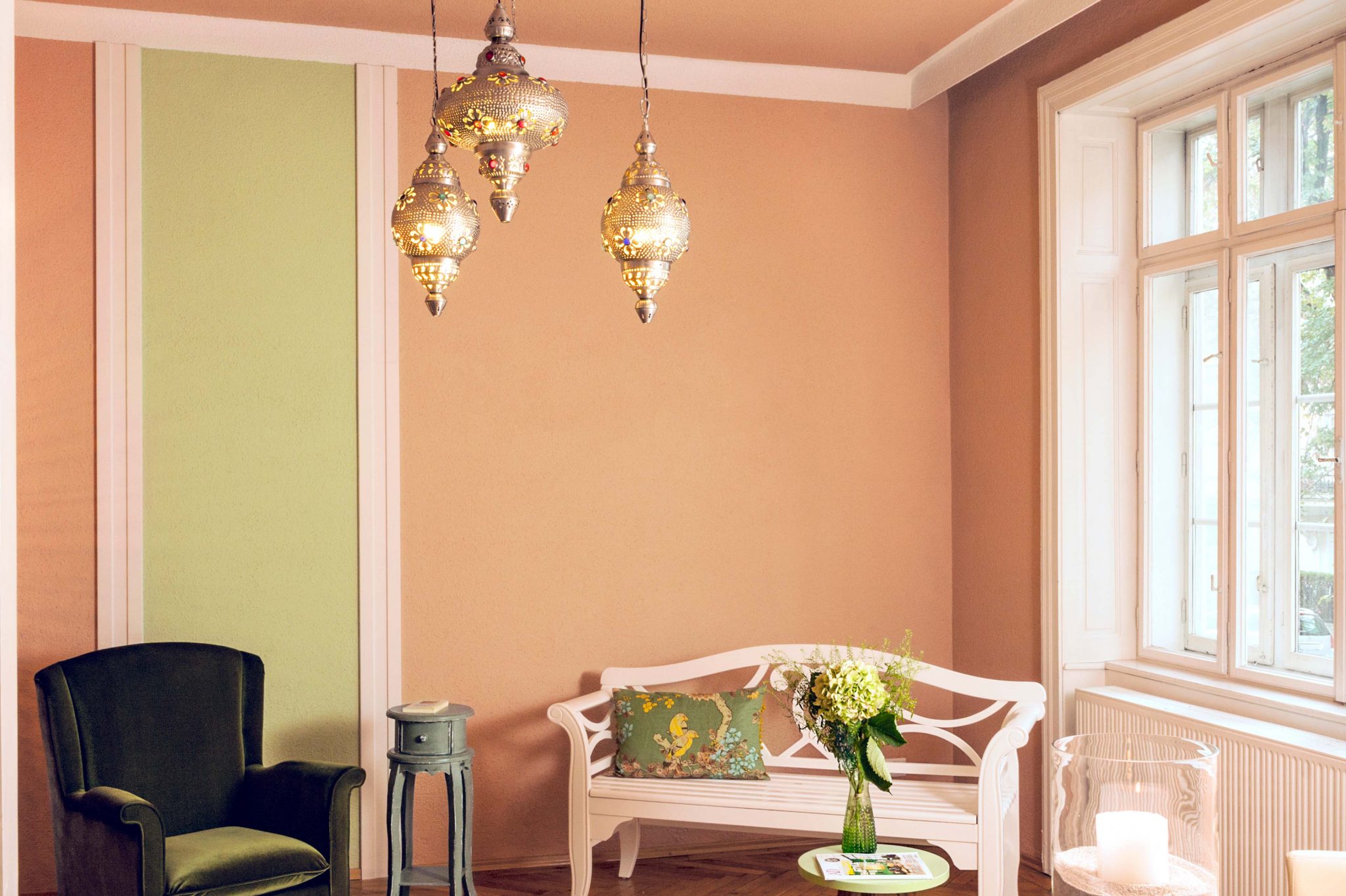 Zimmer eingerichtet mit weißer Bank, grünem Samtsessel, kleinem Tisch mit Blumendekoration und Kerze, orientalische Deckenlampe und Wand in den Farben Cappuccino und lindgrün.