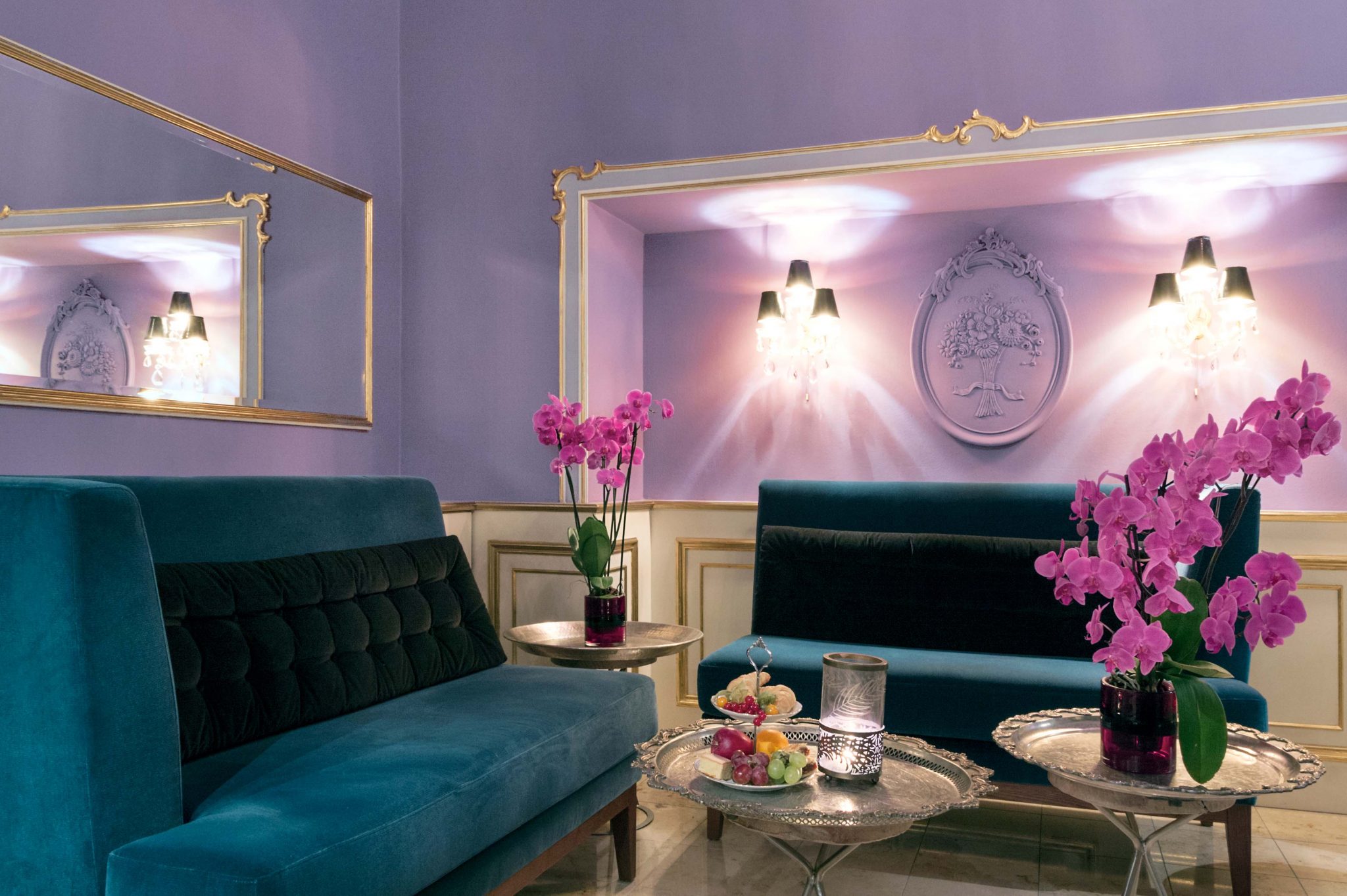 Große, petrolfarbene Samtsofas in flieder-farbenem Raum mit silbernen Tischen und Orchideen