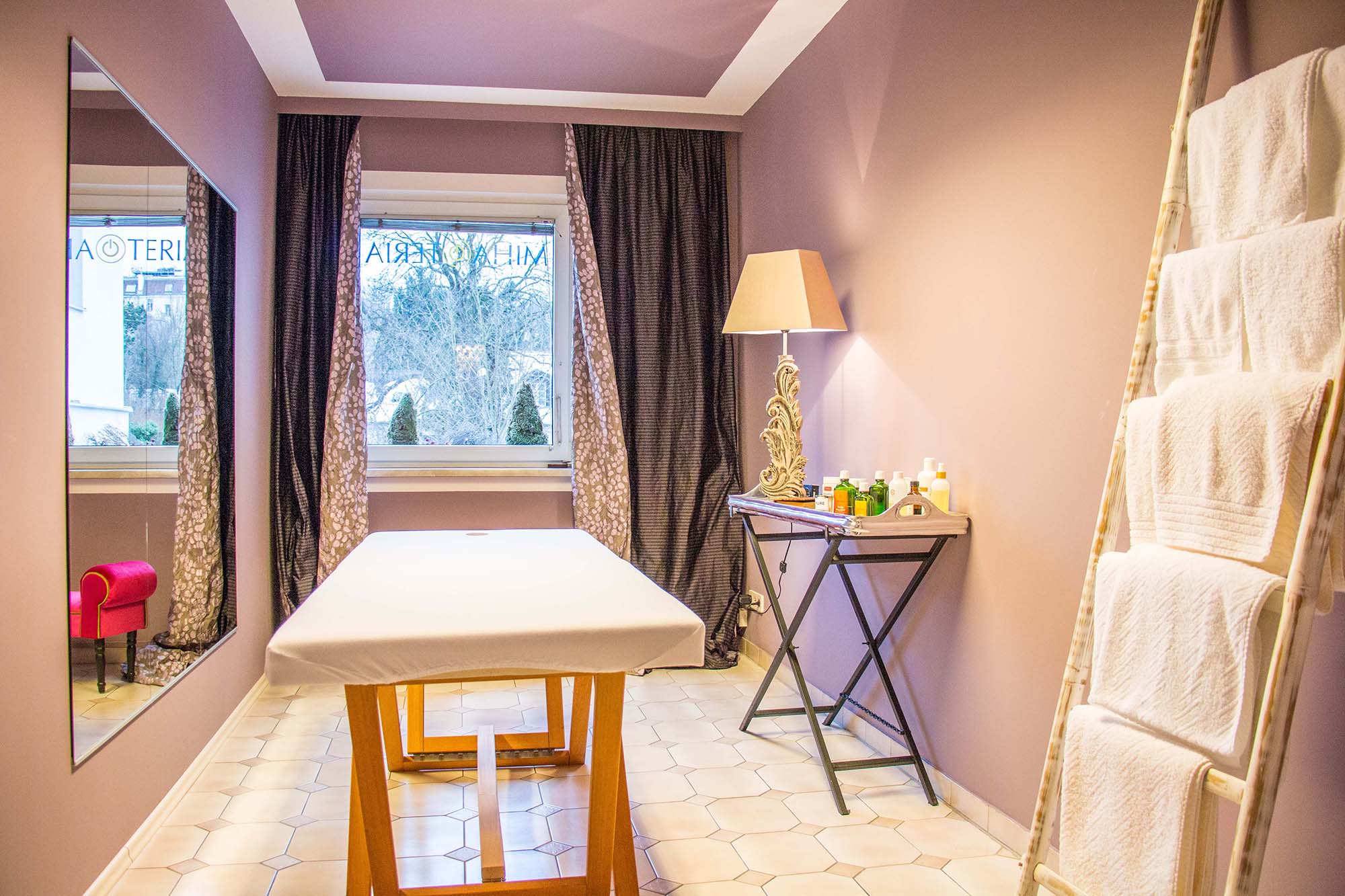 Kleines Zimmer mit pudrig abgetönter lilafarbener Wand, Massageliege, kleinem Sideboard und weißem Handtuchständer aus Holz.
