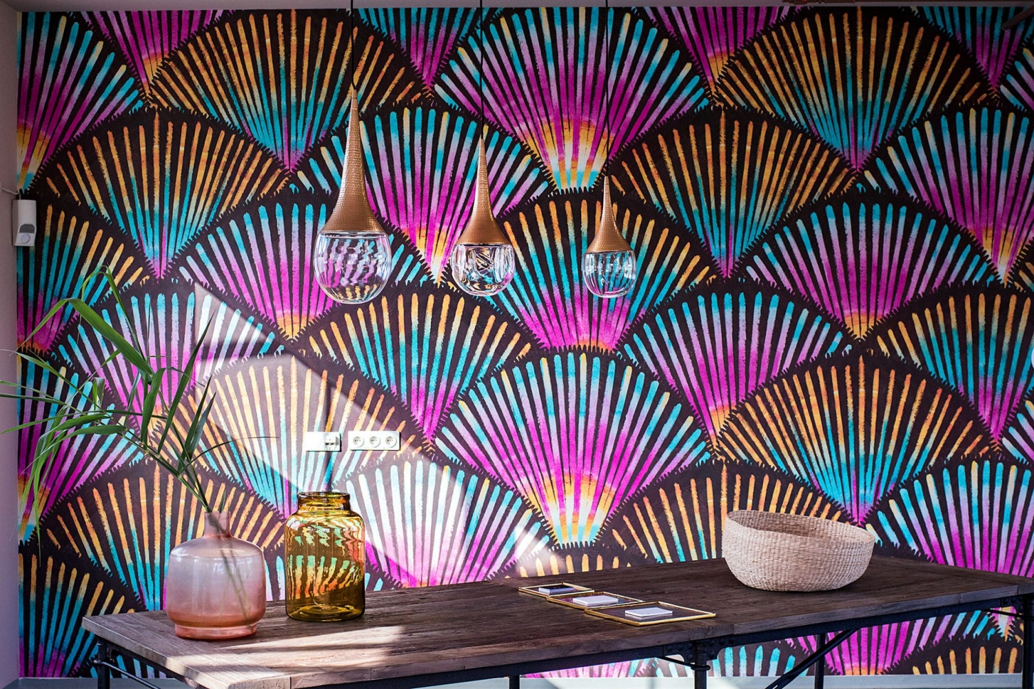 Wallpepper Tapete mit Palmenfächermuster in Schwarz, Gelb, Pink und Blau mit einfallendem Sonnenlicht hinter Holztisch mit kugeligen Hängeleuchten aus Glas und Messing
