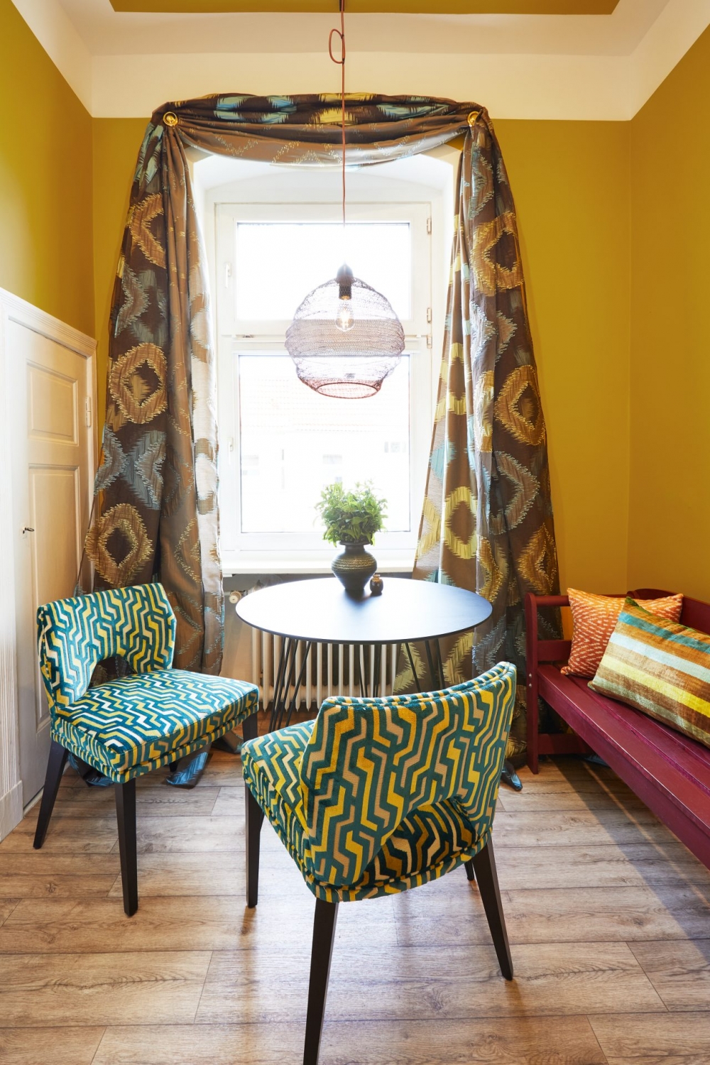 Blick auf Fenster, davor grün-gelb gemusterte Stühle mit farblich abgestimmtem Vorhang, Wandfarbe in Curry und Deckenlampe mit durchsichtigem Schirm