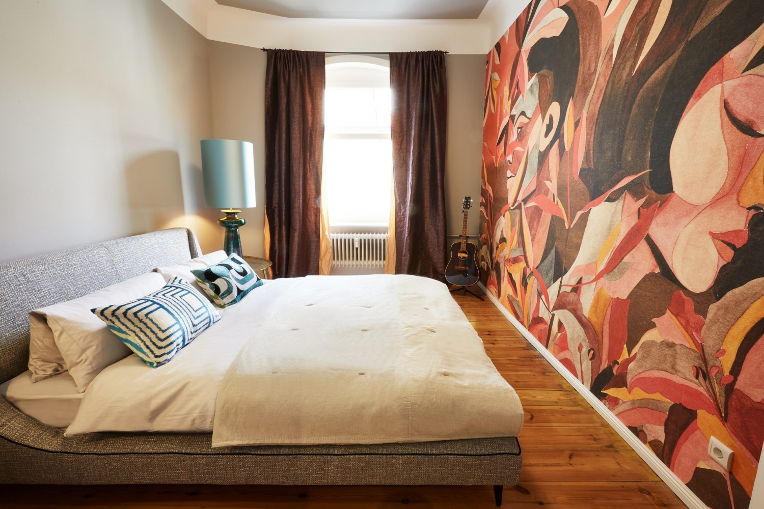Schlafzimmer mit gepolstertem Valentini Bett, Gitarre, blauer großer Lampe auf goldenem Nachttisch und in Herbstfarben gehaltener Tapete mit Männer- und Frauengesicht