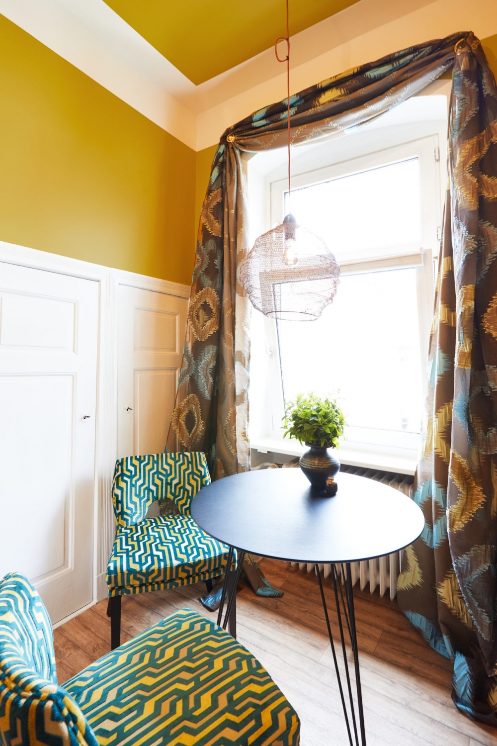 Grün-gelb gemusterte, gepolsterte Stühle mit schwarzem Tisch vor Fenster, mit farblich passenden Vorhängen und curry-farbener Wandfarbe