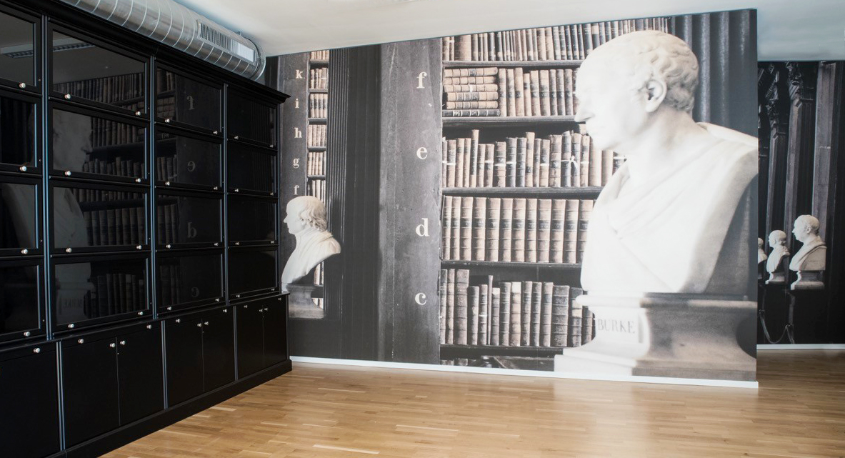 In dunklen Farben gehaltener Raum mit verglaster, schwarzer Bibliothek aus Holz und Tapete mit Motiv von antiken Büchern und Büste