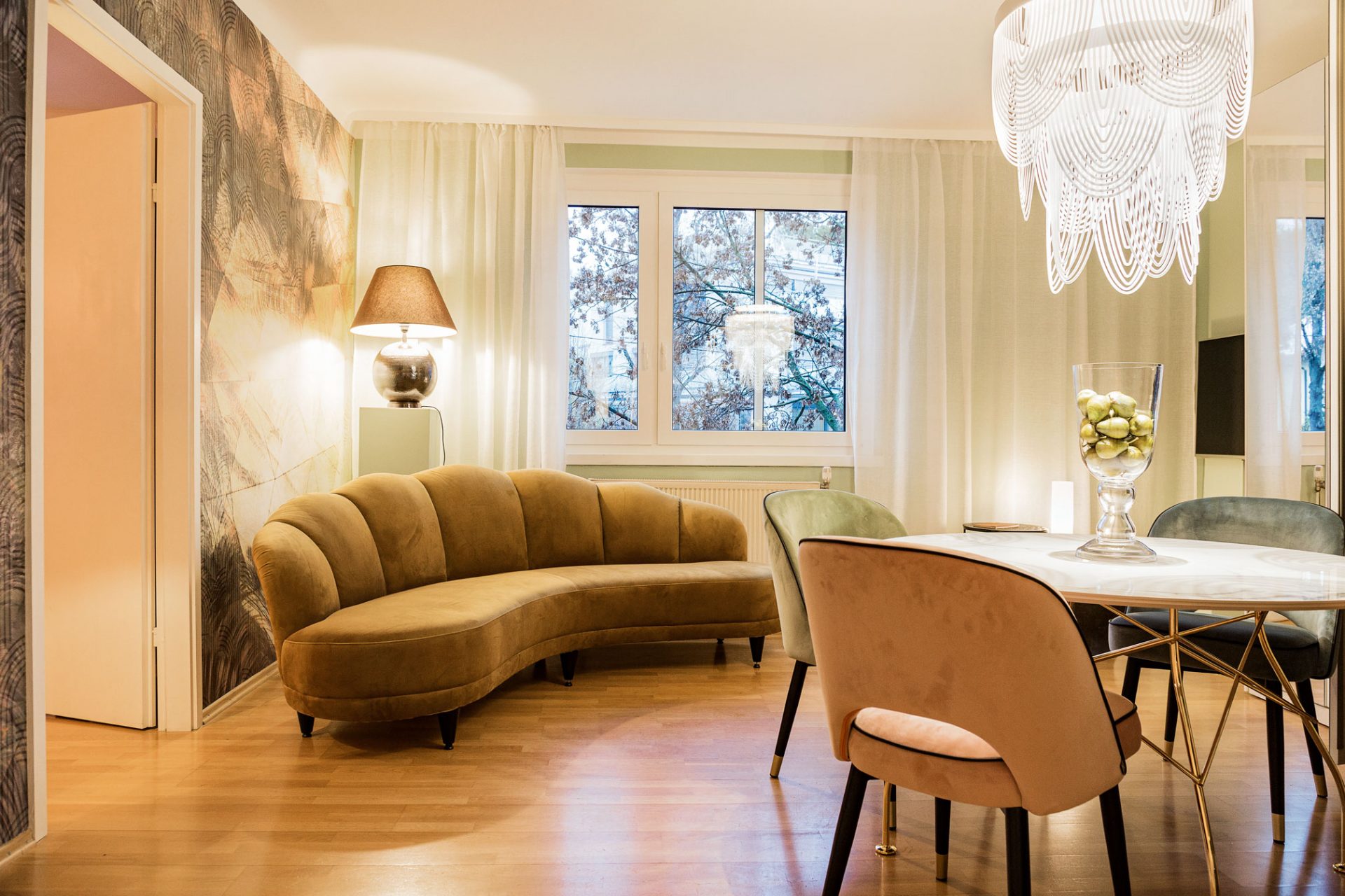 Wohnzimmer mit weißem, modernem Lüster über Marmortisch mit goldenen Beinen, goldfarbenem Sofa, grafisch gemusterter Tapete und pistaziengrüner Wand