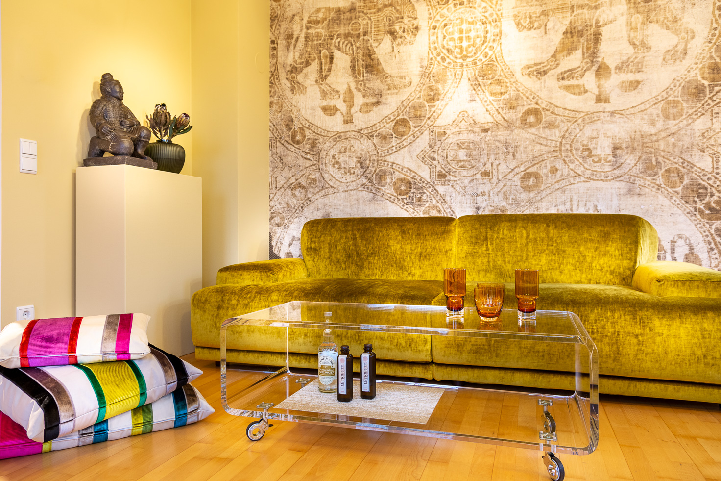 Goldfarbenes, samtiges Sofa von Twils mit Sofatisch aus Acryl, daneben Sitzkissen und Podest mit Terrakotta-Krieger Figur vor byzantinischer Tapete.