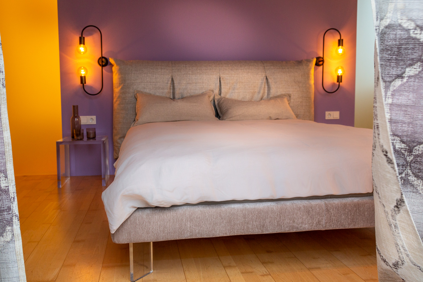 Frontalansicht Bett mit dick gepolstertem Kopfteil und Acrylfüssen, mit Acryl-Nachttisch und eindrucksvollen Wandlampen, Wandfarbe Lila
