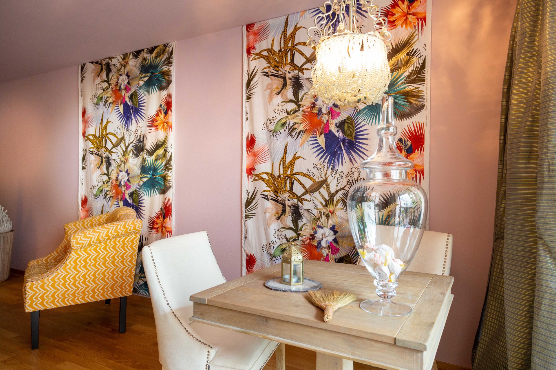 Kleiner Holztisch vor Lacroix-Wandbehang mit verschiedenen blühenden Blumen und Pflanzen.
